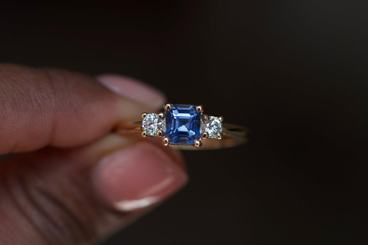 Close up of asscher cut blue sapphire trilogy ring with two round brilliant diamonds in classic yellow gold three stone setting