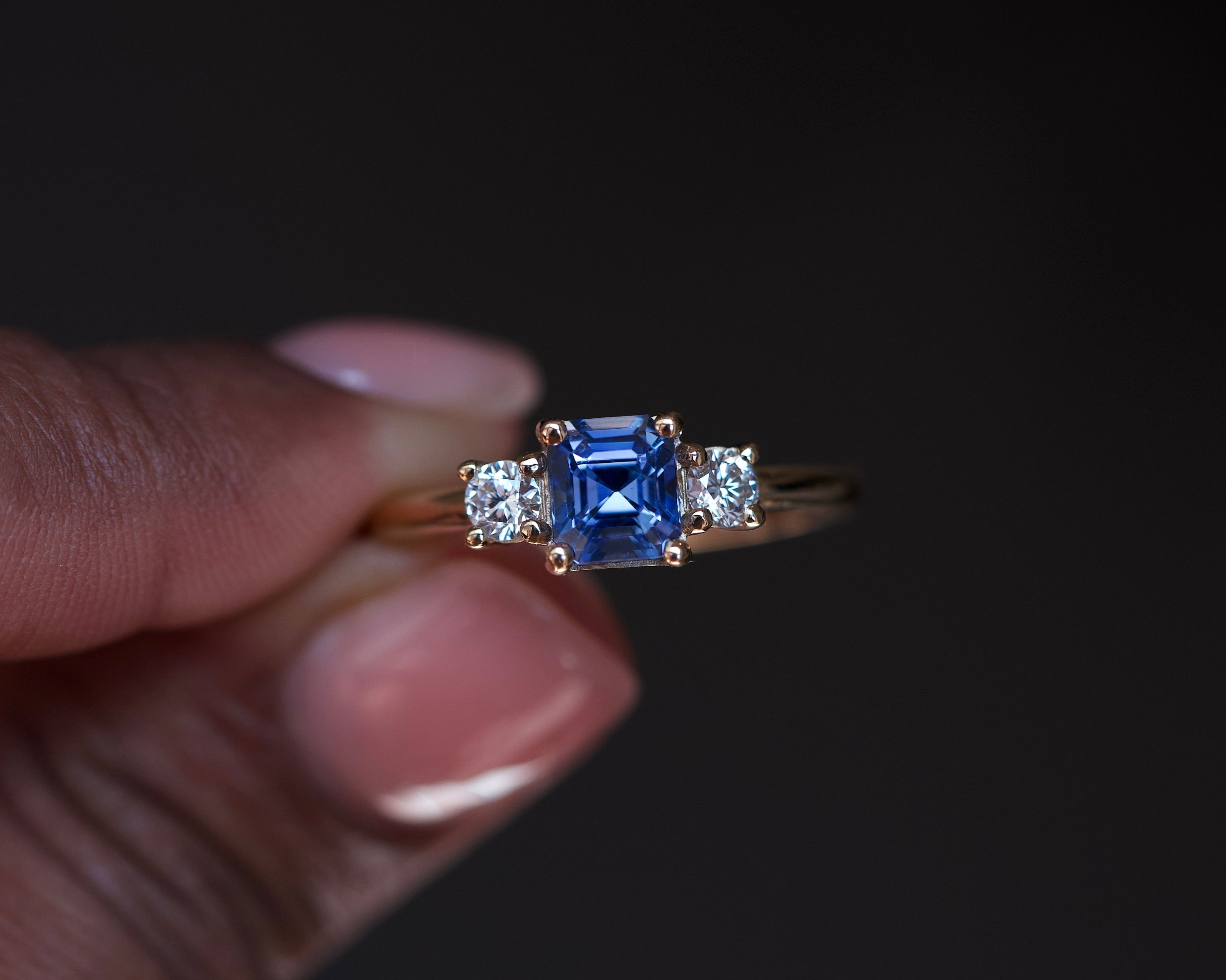 Ceylon blue sapphire trilogy ring with diamond accents in 18k yellow gold, held between fingers against black background showing asscher cut center stone