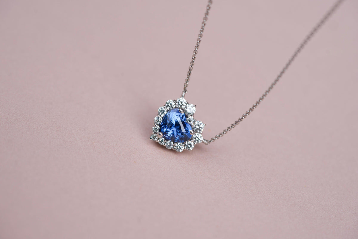 Elegant blue sapphire heart pendant with graduated diamond border against a soft pink backdrop