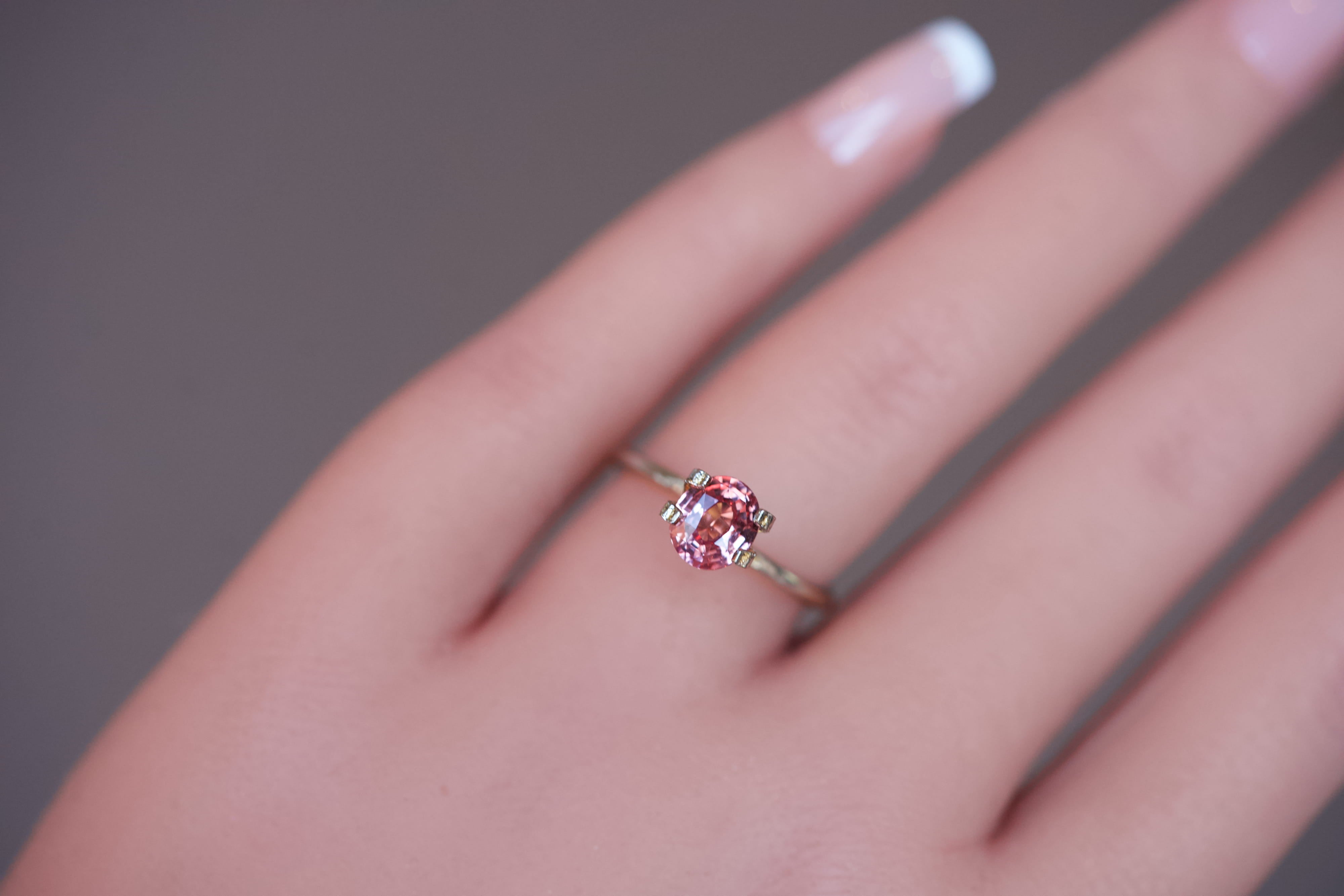 A heated oval Padparadscha sapphire exhibiting pinkish-orange hues, displayed in a gold mock-up ring on a hand.