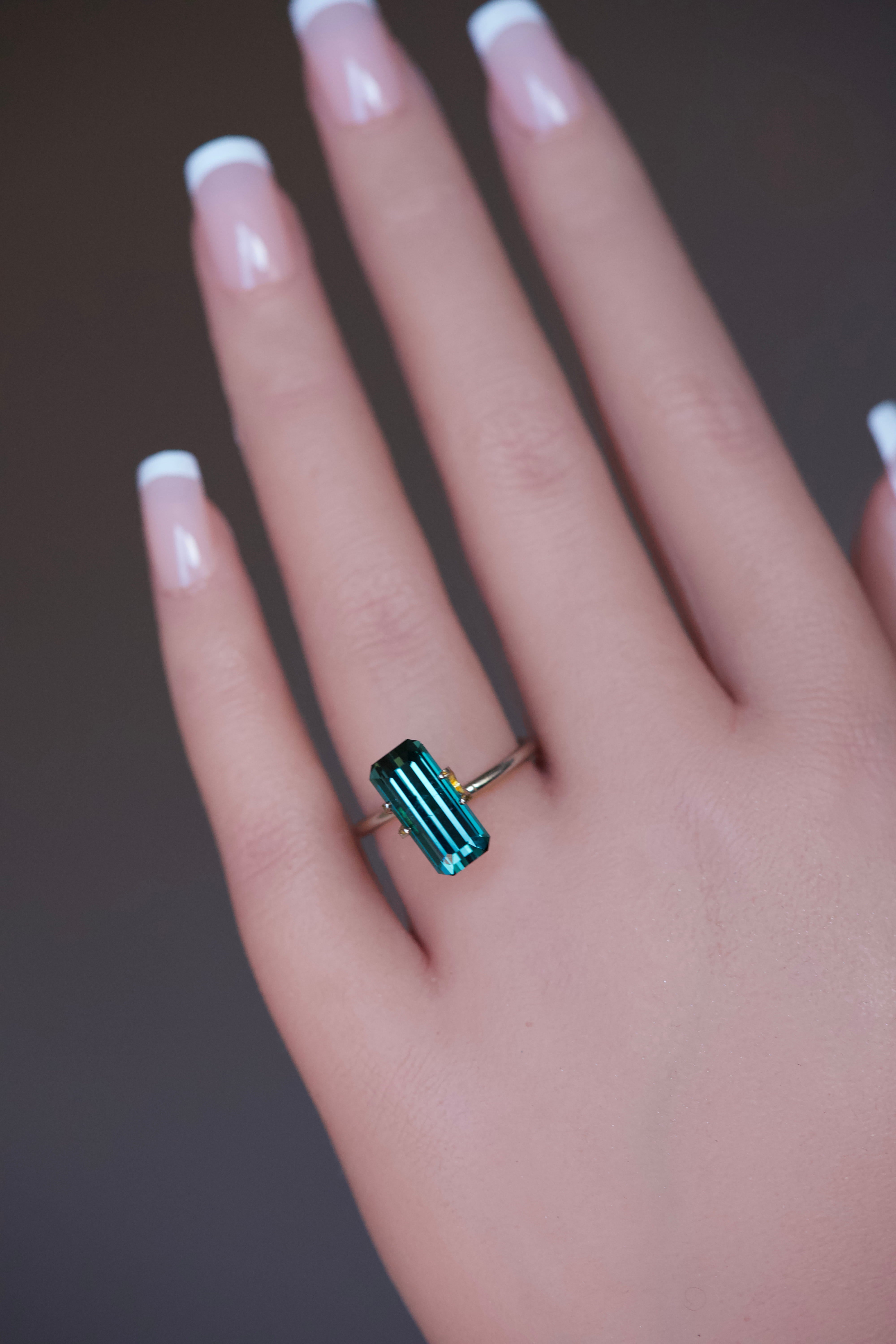 An elongated bluish green tourmaline with a rectangular step cut tried on a hand.