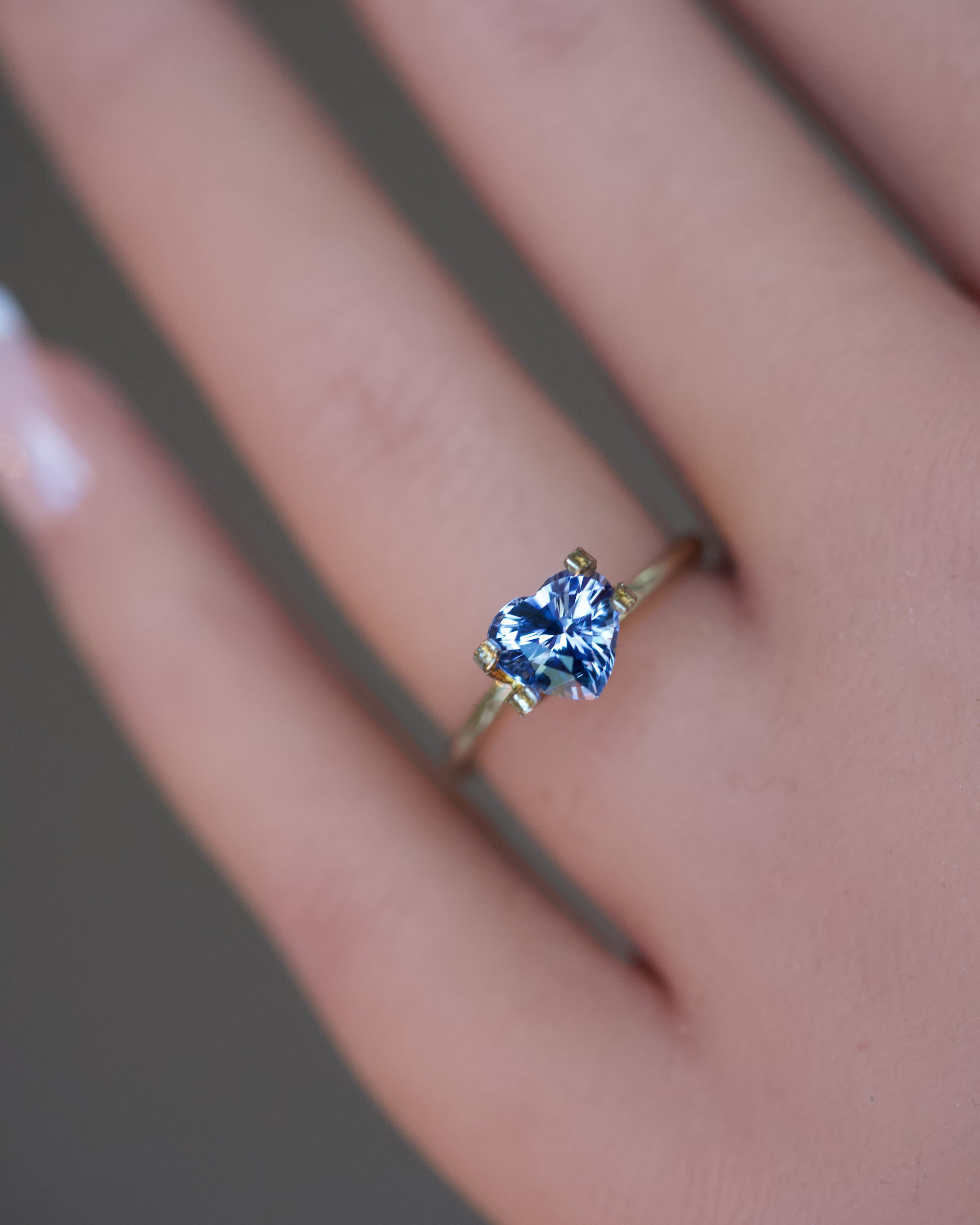 A romantic, unheated bluish-violet heart-shaped sapphire displayed in a temporary gold ring on a finger.