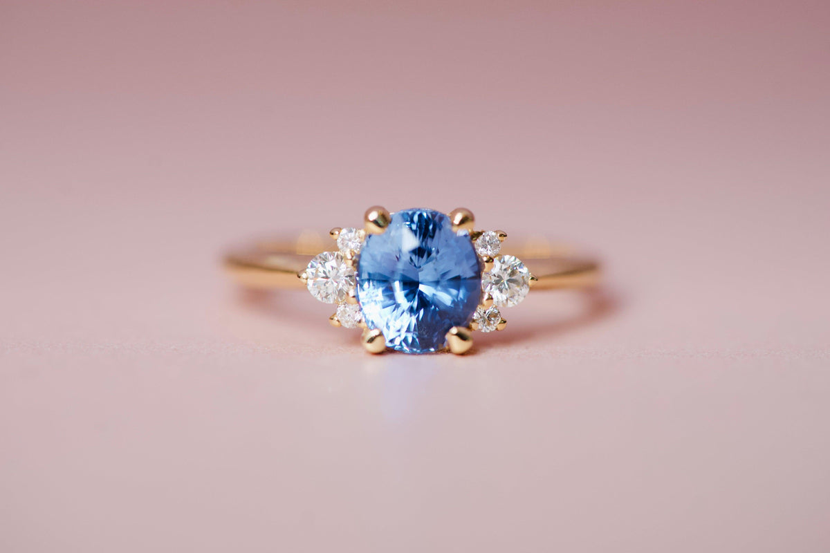 Wide shot of a blue sapphire engagement ring upgrade with small diamond accents on a pink background