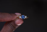 Ceylon blue sapphire trilogy ring with diamond accents in 18k yellow gold, held between fingers against black background showing asscher cut center stone