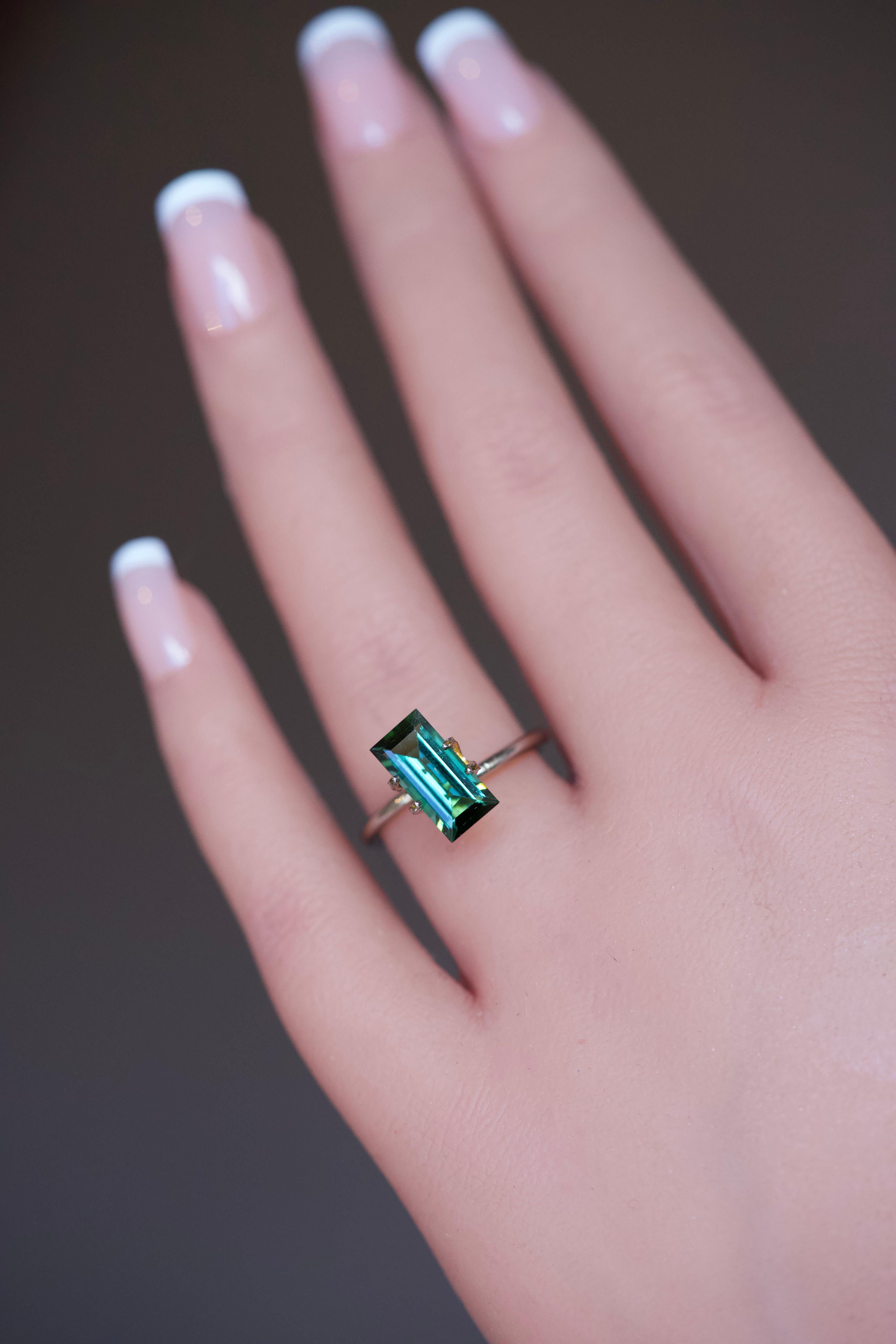 A vibrant 2.85 carat rectangular step-cut green Tourmaline showcased in a temporary gold ring setting on a model's hand.