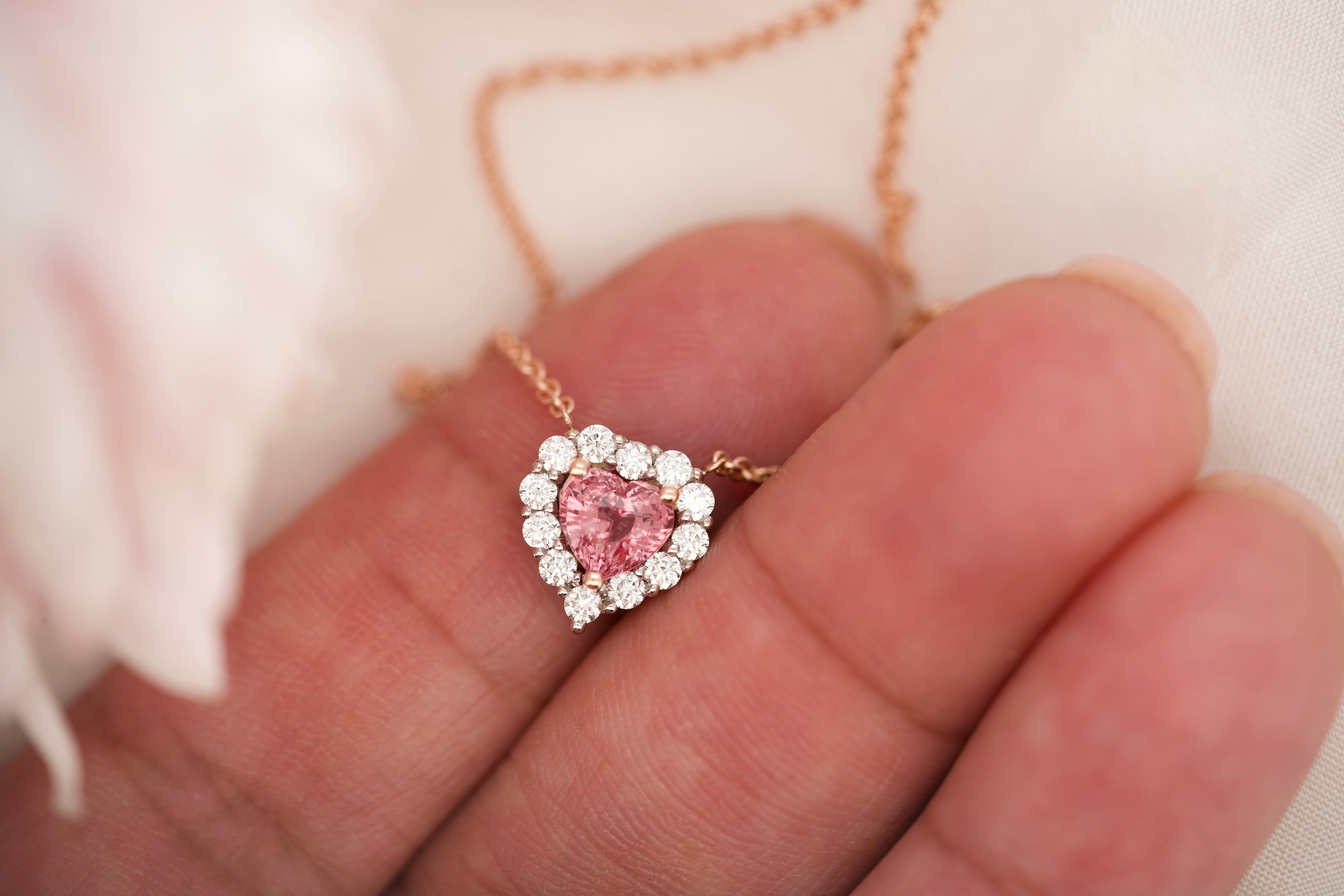 Heart-shaped padparadscha sapphire and diamond necklace resting on a hand.